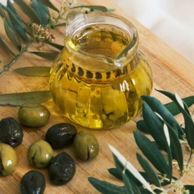 Close-up of fresh olive oil in a glass jar with olives and olive branches on a wooden cutting board.