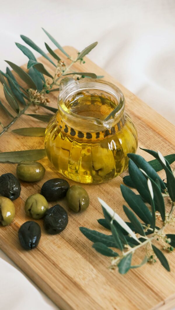 Close-up of fresh olive oil in a glass jar with olives and olive branches on a wooden cutting board.