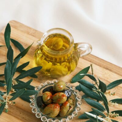 Glass jug of olive oil and a bowl of olives on a wooden cutting board with olive branches.