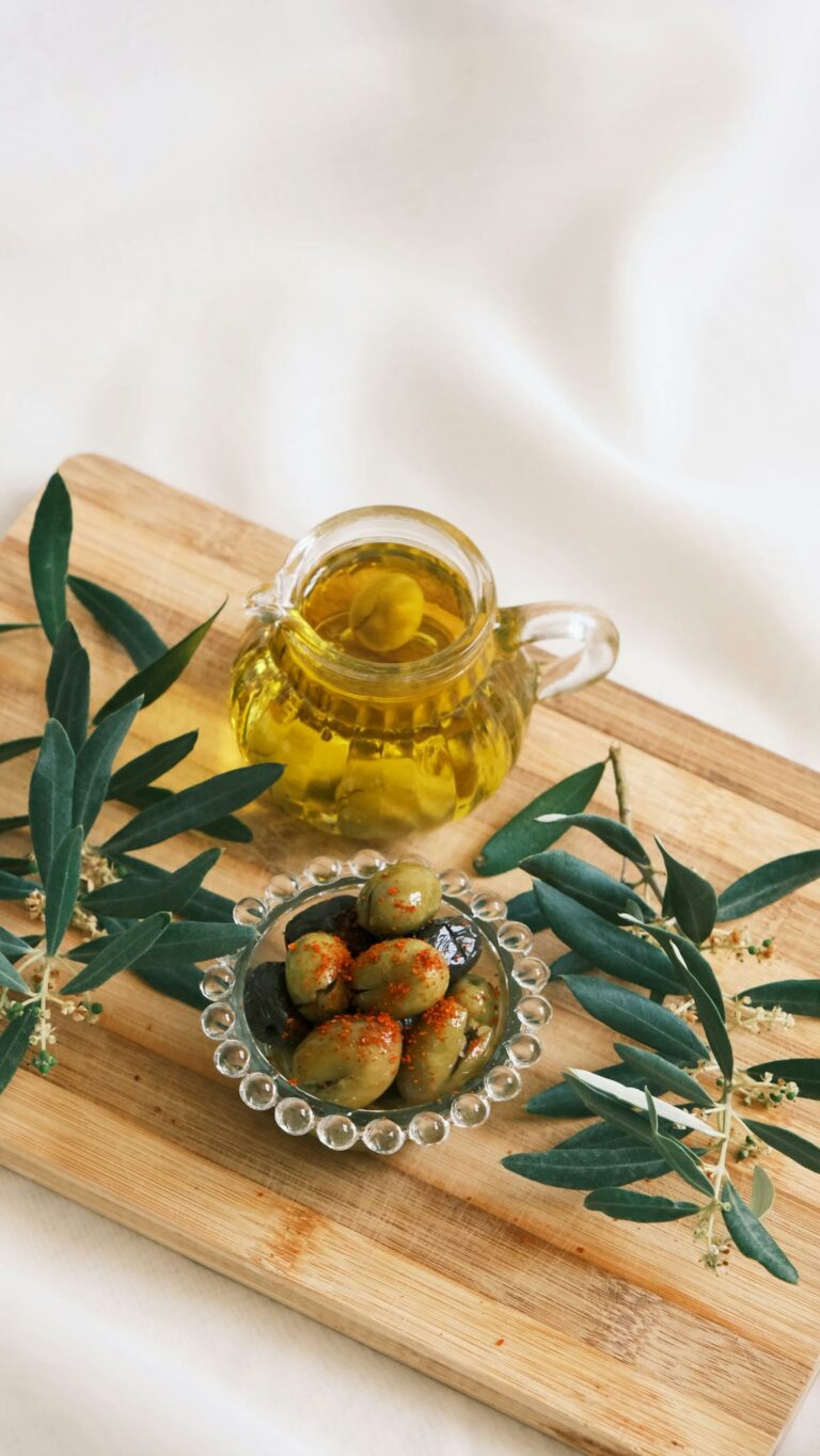 Glass jug of olive oil and a bowl of olives on a wooden cutting board with olive branches.